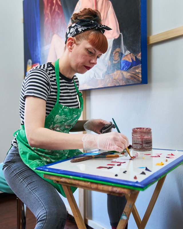 An artist in a stained apron dabs her paintbrush in red oil paint on a glass palette while working in her studio in front of a partially-complete depiction of a recumbent dude.