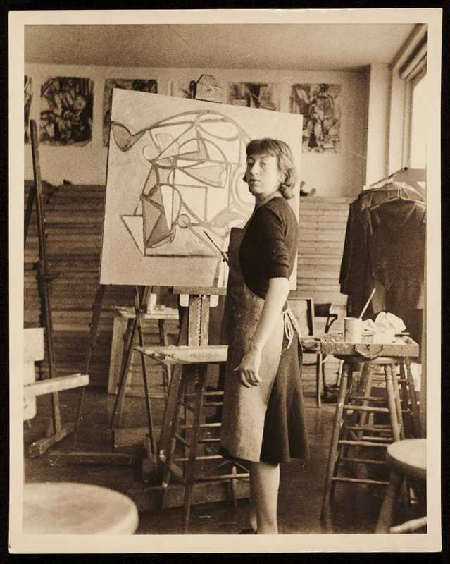 A monochromatic photograph of Lee Krasner standing in front of an abstract painting with raised eyebrows and paintbrush in hand. Image caption: Lee Krasner in Hans Hoffman’s studio, early 1940s. Photo ©Robert E. Mates and Paul Katz. Lee Krasner artwork ©Pollock-Krasner Foundation/ARS. Image courtesy of the Pollock-Krasner Foundation.